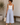 Woman wearing a white tank top and light blue pants, holding a woven bag indoors.