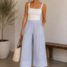 Woman wearing a white tank top and light blue pants, holding a woven bag indoors.