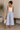 Woman wearing a white tank top and light blue pants, holding a woven bag indoors.