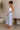 Woman wearing a white tank top and light blue pants standing in a room with a plant and wooden bench.