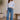 Woman wearing a light blue shirt and jeans standing in a room with a plant and wooden bench.