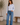 Woman wearing a light blue shirt and jeans standing in a room with a plant and wooden bench.