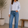 Woman wearing a light blue shirt and jeans standing in a room with a plant and wooden bench.