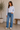 Woman wearing a light blue shirt and jeans standing in a room with a plant and wooden bench.