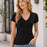 A woman wearing a black short-sleeve V-neck t-shirt and light blue jeans, standing outdoors in front of a white stucco building with a window and wall lantern.
