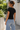 A woman wearing a fitted black short-sleeve t-shirt and light-wash blue jeans, photographed from the back in an outdoor walkway with white walls and greenery.