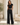 A woman stands indoors wearing a black short-sleeve crop top and black wide-leg pants with white side stripes, paired with white sneakers. The room has a light neutral wall, a potted plant on the left, and a cream-colored armchair on the right.