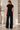 A woman shown from the back wearing a black short-sleeve t-shirt and matching black wide-leg pants with white side stripes, standing in a bright indoor room.