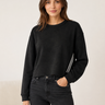 A woman modeling a black cropped crewneck sweatshirt with white stripes down the sleeves, paired with dark gray jeans, against a neutral cream background.