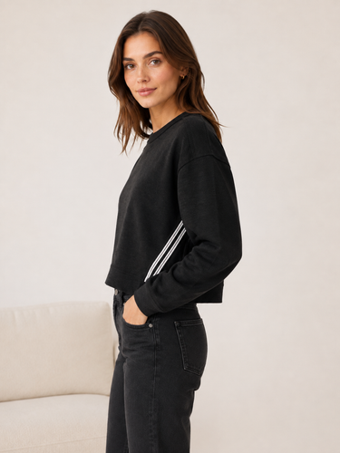 A woman wearing a black cropped sweatshirt with white stripes on the sleeves, paired with black jeans, standing in a studio setting.