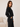 A woman wearing a black cropped long-sleeve sweatshirt with white stripes on the sleeve, paired with black jeans, standing in a minimalist room with a light beige sofa.