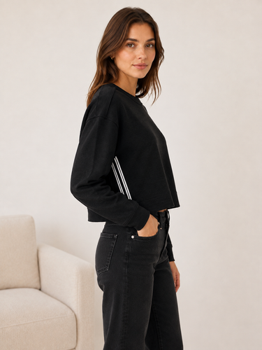 A woman wearing a black cropped long-sleeve sweatshirt with white stripes on the sleeve, paired with black jeans, standing in a minimalist room with a light beige sofa.