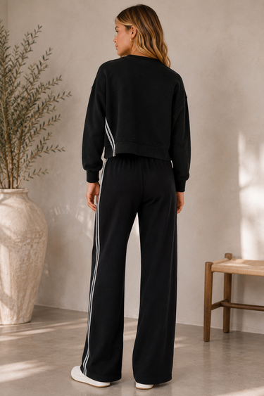 A woman stands with her back to the camera wearing a matching black tracksuit with white side stripes, featuring a cropped sweatshirt and wide-leg pants, in a minimalist indoor setting.