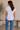 Woman wearing a white striped shirt and blue jeans standing in a room with a plant and wooden bench.