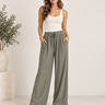 A woman modeling a pair of wide-leg, high-waisted sage green pants with a drawstring waist and side pockets, paired with a white tank top and brown sandals in a bright studio setting.