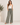 A woman modeling a pair of wide-leg, high-waisted sage green pants with a drawstring waist and side pockets, paired with a white tank top and brown sandals in a bright studio setting.
