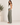 A woman stands in profile wearing wide-leg sage green drawstring pants and a white tank top in a bright, neutral indoor setting with a potted plant on the left.