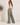 A woman wearing high-waisted, wide-leg sage green pants and a white tank top, standing in a minimalist studio setting with a potted plant on the left.