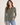 A woman modeling a long-sleeve button-up shirt in olive green, paired with blue jeans, against a plain light beige studio background.