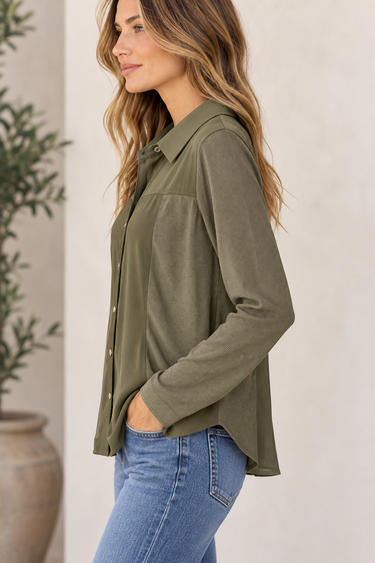 A woman wearing an olive green long-sleeve button-up shirt and blue jeans, shown in a side profile against a light beige wall with a potted plant in the background.