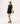 A model facing backwards wearing a black dress under a black lace long-sleeve tunic cover-up with scalloped pattern, lace-up V-neck and tassel ties.