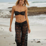 A woman wearing black lace sheer cover up pants over a black bikini on a beach.