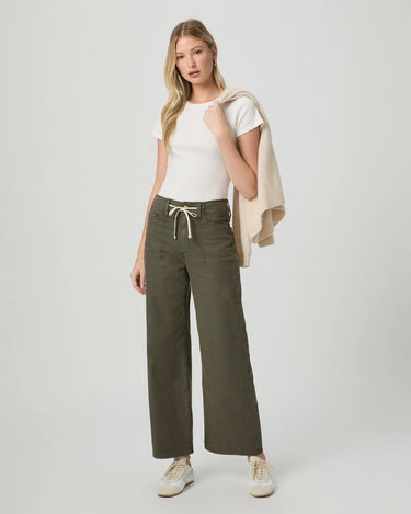 A woman modeling olive green wide-leg pants with a cream drawstring, styled with a white t-shirt, a beige cardigan draped over her shoulder, and white sneakers, set against a plain light gray studio background.