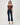 A woman modeling an off-the-shoulder brown top and dark blue cropped flare jeans with brown strappy heels, shot against a light gray studio background.