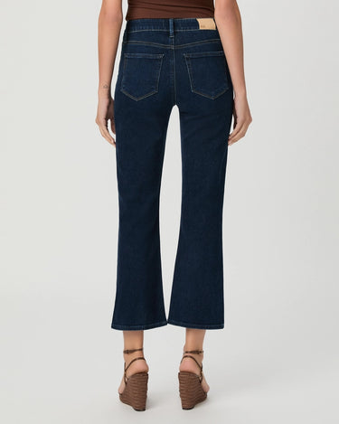 A person shown from the back wearing dark blue cropped flare jeans with a brown top and brown wedge sandals against a light gray studio background.