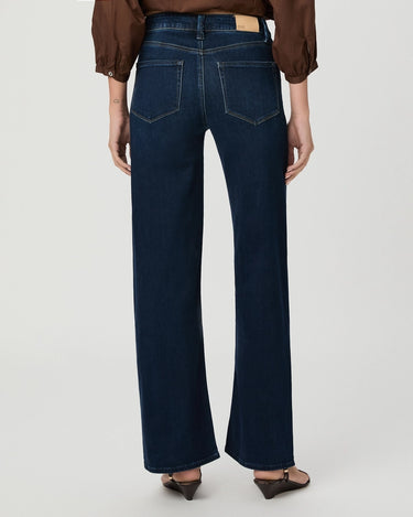 Rear view of a model wearing dark blue wide-leg jeans with a brown blouse and brown shoes, against a light gray studio background.
