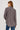 A woman shown from the back wearing a long-sleeve button-up shirt with vertical blue, brown, and white stripes, paired with blue jeans, against a plain light gray studio background.