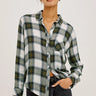 A woman modeling a long-sleeve plaid button-up shirt in white, green, and gray, paired with dark jeans and sunglasses, against a plain light background.