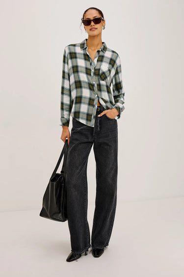 Person wearing a green and black plaid shirt with black jeans, holding a black bag on a white background