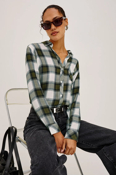 A woman wearing a green, white, and black plaid button-down shirt, dark gray jeans, and sunglasses, sitting on a clear chair against a light gray studio background.