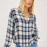 A woman wearing a long-sleeve button-up plaid shirt in blue, white, and navy, paired with light-wash blue jeans, photographed against a light gray studio background.