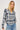 A woman wearing a long-sleeve button-up plaid shirt in blue, white, and navy, paired with light-wash blue jeans, photographed against a light gray studio background.
