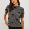 A woman wearing a short sleeve charcoal sweater with velvet black bows and a black midi skirt on a white background.