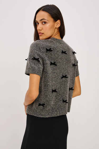 A woman wearing a short sleeve charcoal sweater with velvet black bows and a black midi skirt on a white background.