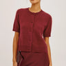 A model wearing a short-sleeve button-front knit cardigan in a deep burgundy red, paired with a matching skirt and sunglasses, against a plain light gray studio background.