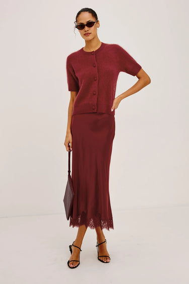 A model wearing a matching burgundy knit short-sleeve cardigan and midi skirt set, styled with sunglasses, a dark handbag, and black strappy heels against a plain white studio background.