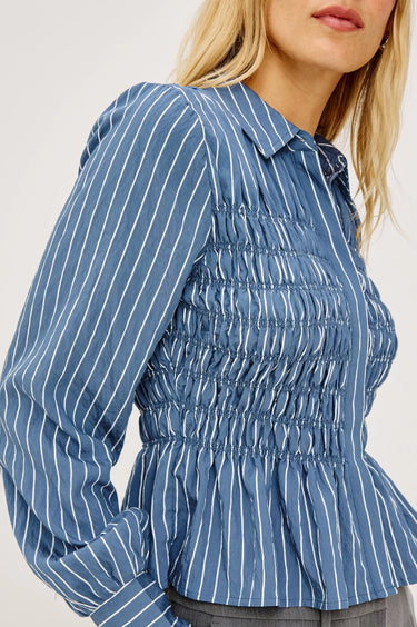 A close-up of a woman wearing a blue long-sleeve blouse with white vertical pinstripes and a smocked bodice, cropped from the mouth down against a plain light background.