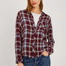 A woman modeling a long-sleeve button-front plaid shirt in burgundy, gray, and white, paired with blue jeans against a light gray studio background.