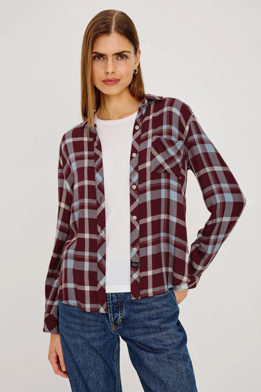 A woman modeling a long-sleeve button-up plaid shirt in burgundy, gray, and white, worn open over a white t-shirt and blue jeans, against a light gray studio background.
