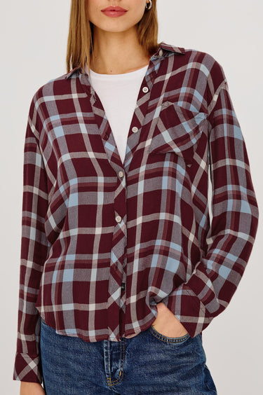 A woman wearing a long-sleeve button-up plaid shirt in burgundy, light blue, and white, paired with a white t-shirt and blue jeans.