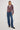 A woman modeling a long-sleeve button-up plaid shirt in burgundy, gray, and white, paired with wide-leg blue jeans and dark brown shoes, against a plain white studio background.