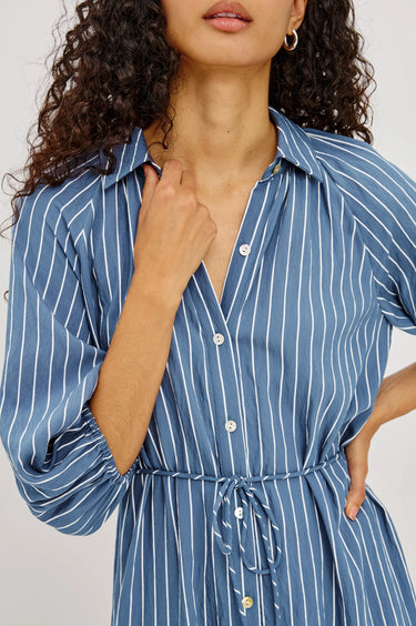A close-up of a model wearing a blue and white vertically striped shirt dress with a collar, button front, and drawstring waist.