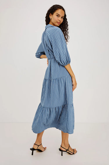 A woman modeling a blue and white vertically striped midi dress with puff sleeves and a tiered skirt, shown from a three-quarter back angle against a plain white studio background.