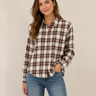 A woman modeling a long-sleeve button-up plaid shirt in cream, brown, and black, paired with blue jeans, against a plain beige studio background.