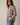 A woman wearing a long-sleeve plaid button-up shirt in cream, brown, and beige tones, paired with blue jeans, photographed against a plain light beige studio background.