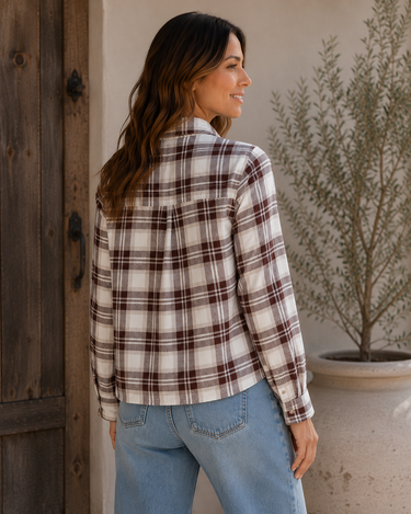 A woman wearing a brown and cream plaid flannel shirt and light blue jeans, standing in profile next to a rustic wooden door with a potted plant in the background.